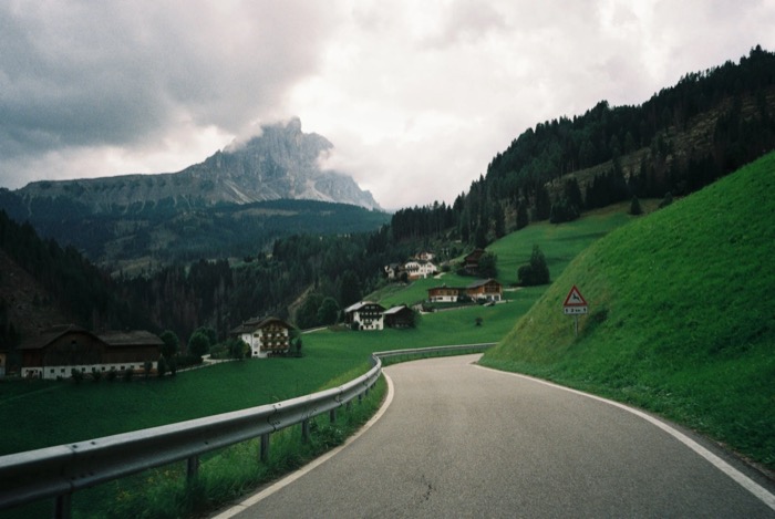 Empty mountain road curving past green pastures and scattered houses toward a cloud-wrapped peak