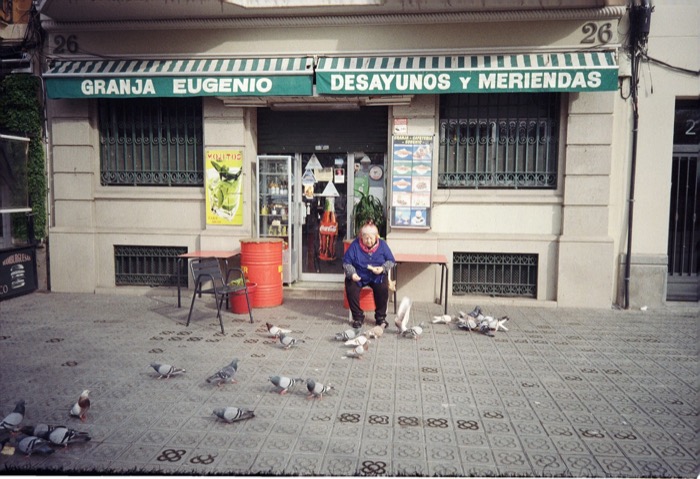 Woman feeding pigeons outside a cafe with a striped awning reading Granja Eugenio, Desayunos y Meriendas
