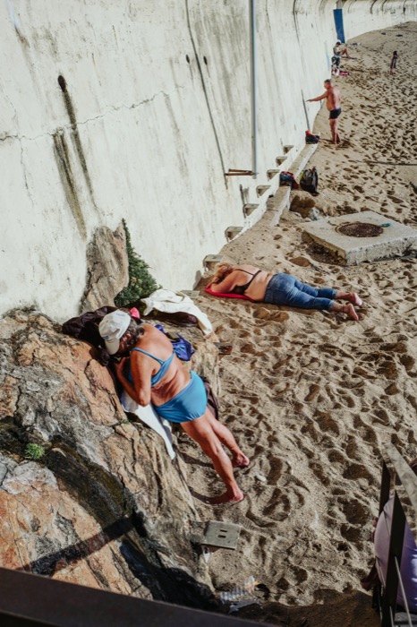 Sunbathers stretched out on sand and rock beside a high concrete sea wall, seen from above