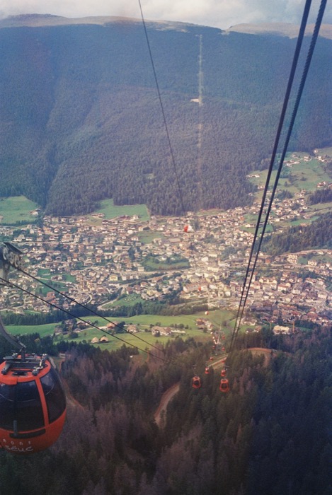 View from a cable car down over a forested valley to an alpine town, red gondolas on the wires