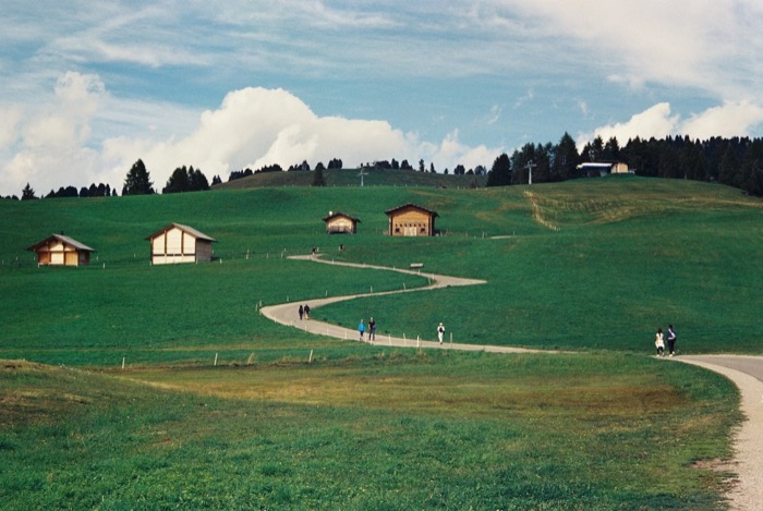 Walkers on a path curving through green alpine meadow past wooden hay barns below a wooded ridge