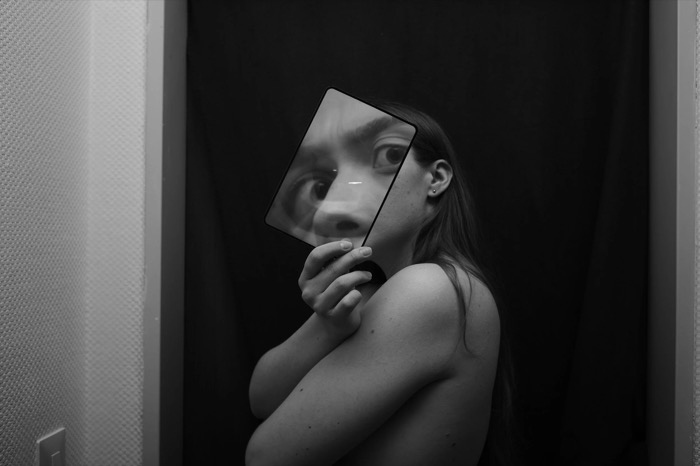 Woman holding a small square mirror over her face, one enlarged eye staring out of the reflection