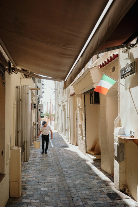 Man carrying a bucket down a cobbled lane under awnings, an Italian flag on the wall
