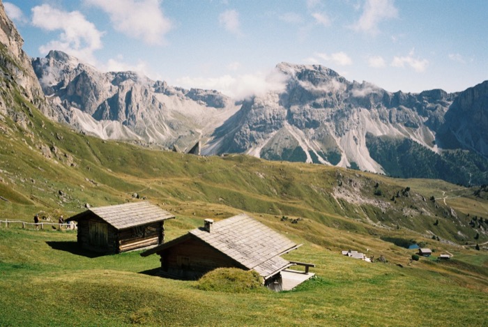Two shingled wooden huts on an alpine meadow below jagged limestone peaks