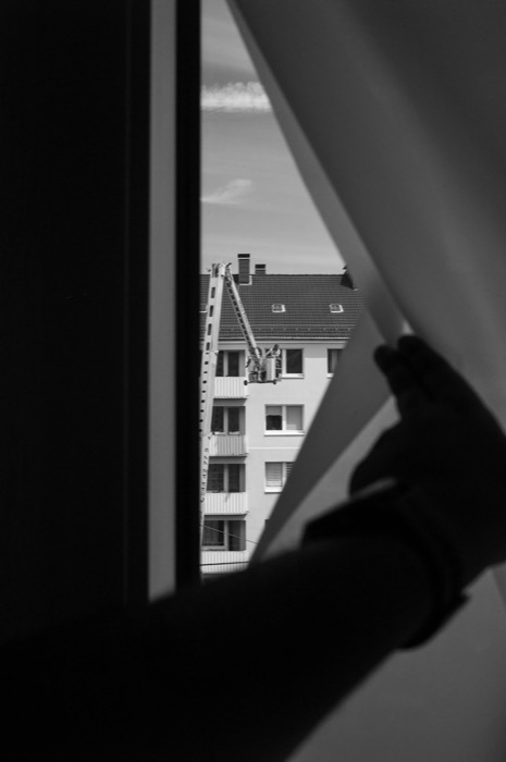 Hand drawing back a curtain on a window; a worker rides a ladder lift up an apartment block outside