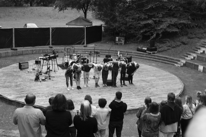 Band lined up bowing on a round open-air stage as the standing audience applauds from below