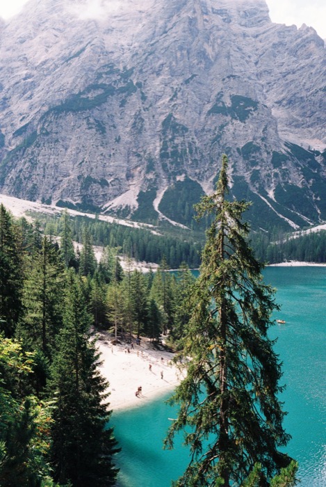 Turquoise mountain lake with bathers on a pale shore, framed by spruce below a huge limestone wall