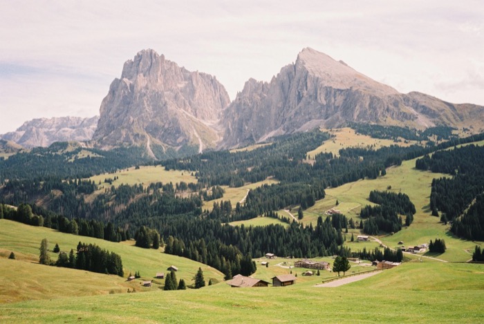 Broad meadow and forest below two massive rock massifs rising over a valley of huts