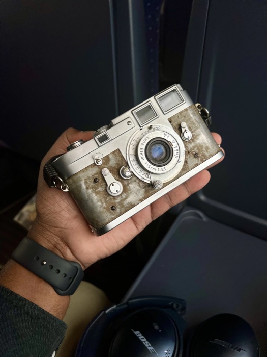Hand holding a battered chrome rangefinder with a collapsible 5cm Elmar lens on an aeroplane seat