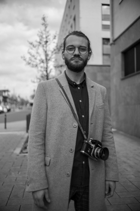 Bearded man in round glasses and a pale overcoat stands on a pavement, a film SLR slung at his hip