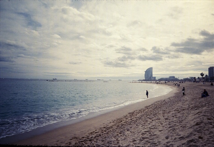 Wide empty beach curving toward a sail-shaped seafront hotel, one figure standing in the shallows
