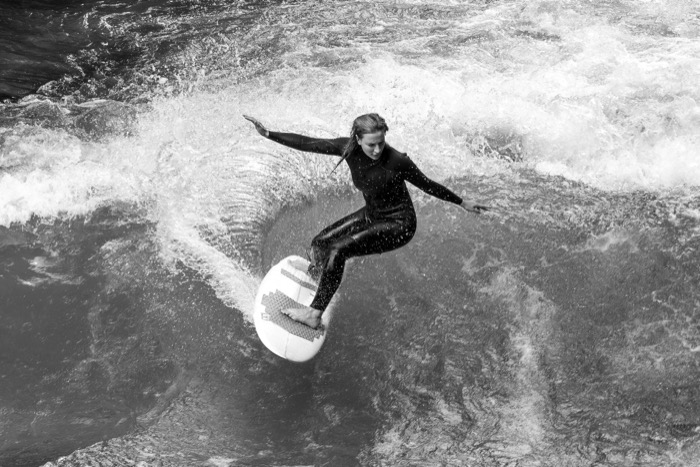 Woman in a wetsuit carving a turn on a standing river wave, arms out for balance amid whitewater