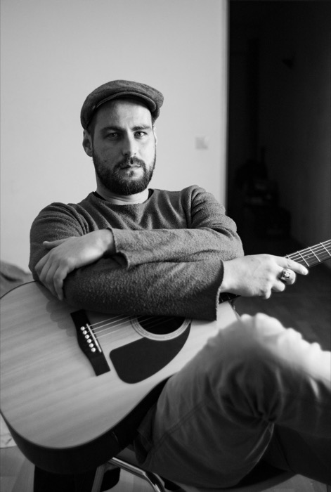 Man in a flat cap rests his crossed arms on an acoustic guitar, looking directly into the lens