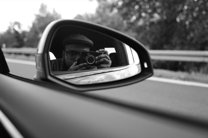 Man in a flat cap raising a rangefinder to his eye, reflected in a car wing mirror on a motorway