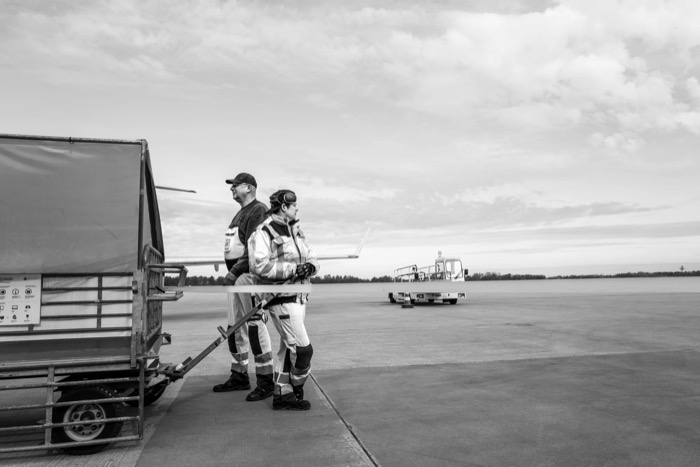 Two ground crew in high-visibility overalls standing by a baggage cart on an empty airport apron