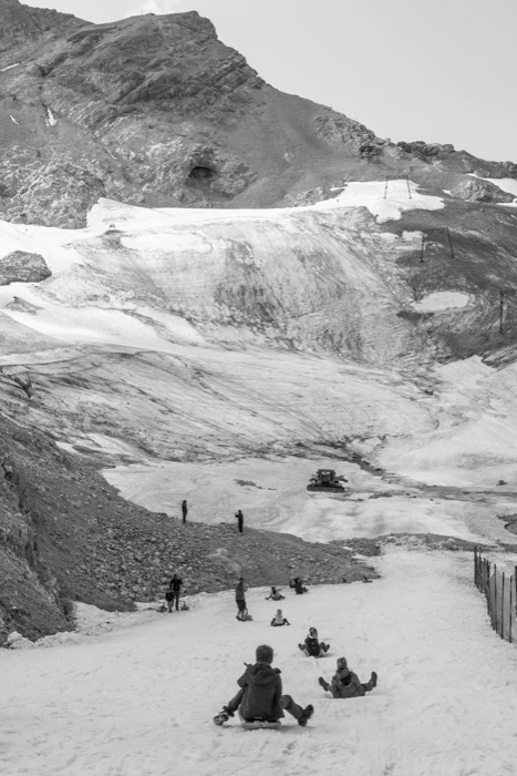 People sledging down a summer snowfield below bare rocky peaks and a shrinking glacier