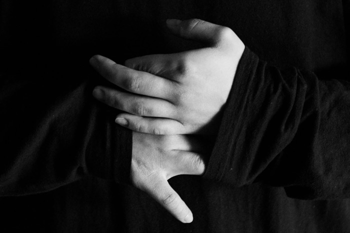 Pale hands folded against dark clothing, the only light in an otherwise black frame