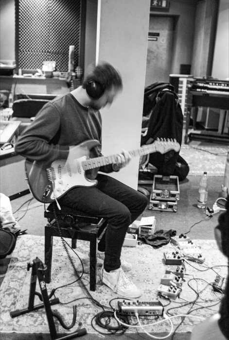 Guitarist in headphones playing a Stratocaster in a studio, pedalboard and cables spread over a rug