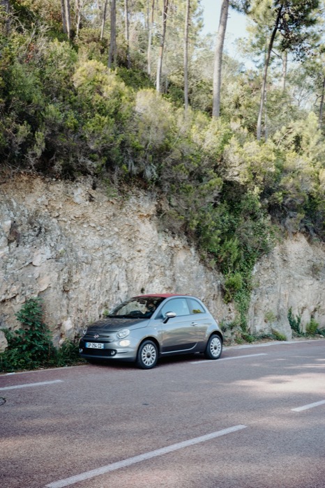 Small grey convertible parked against a rock cutting on a wooded coastal road