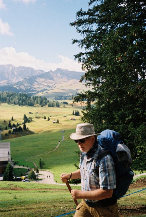 Hiker in a sun hat, sunglasses and heavy blue pack climbing a meadow, mountain valley spread out behind