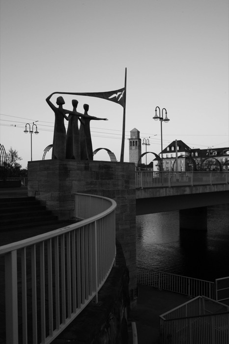 Bronze figures holding a flag on a riverside bridge monument at dusk, curved railing leading in