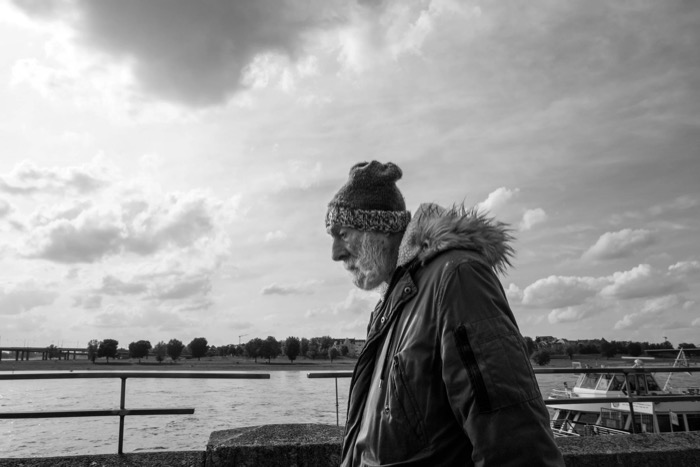 Bearded older man in a bobble hat and fur-trimmed parka walking a river promenade under towering clouds
