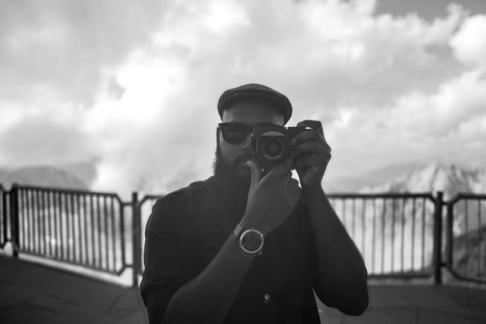 Bearded man in a flat cap and sunglasses raising a rangefinder to his eye on a mountain viewing terrace