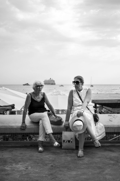 Two women in sunglasses sitting on a seafront wall in conversation, a cruise ship anchored behind them