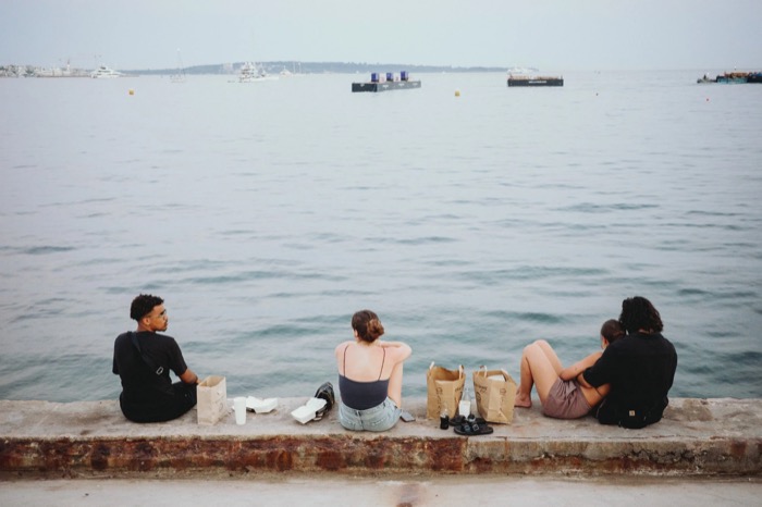 Four young people sitting on a harbour wall with takeaway bags, facing calm water and moored boats