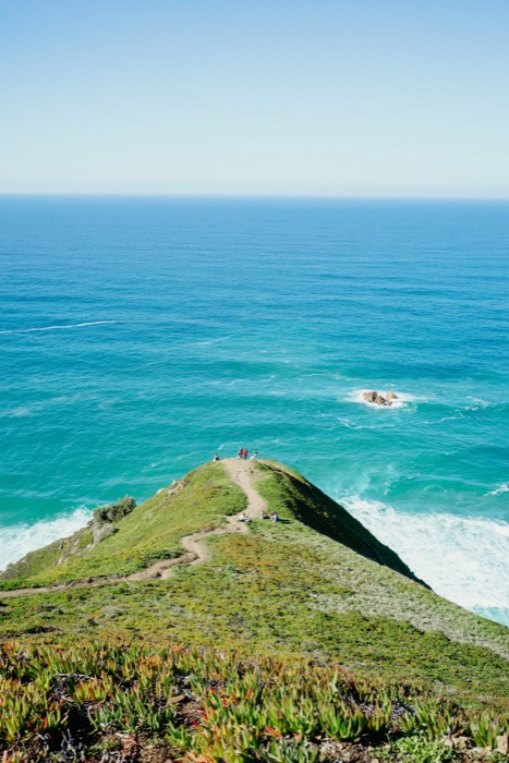 Green headland ridge with a footpath running out to a point above turquoise ocean, tiny figures at the tip