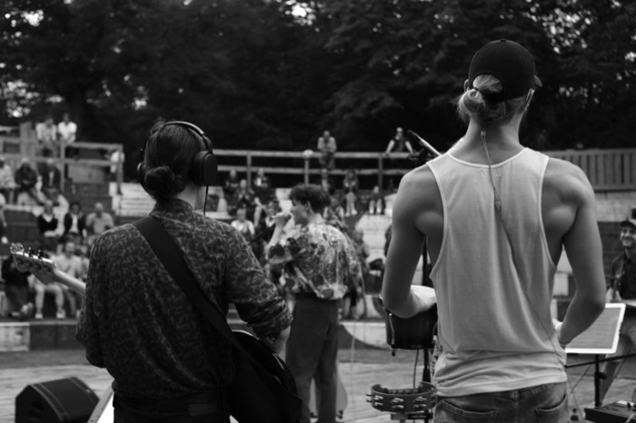 Two musicians seen from behind on stage, one in headphones with a guitar, facing an outdoor amphitheatre crowd