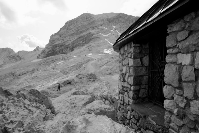 Stone hut at the edge of a bare rocky summit plateau, a lone walker crossing the scree