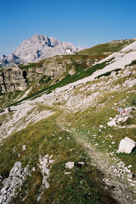 Stony mountain path leading to a small white refuge hut, a broad rock massif rising beyond