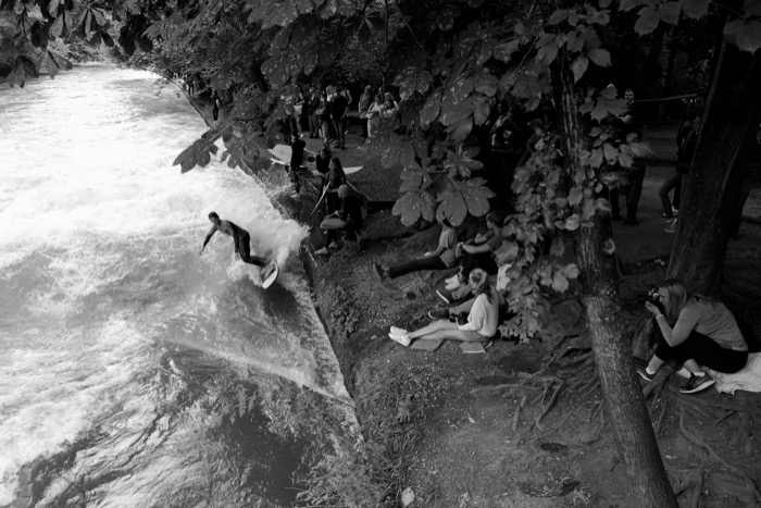 Surfer riding a standing river wave while spectators watch and photograph from the shaded bank