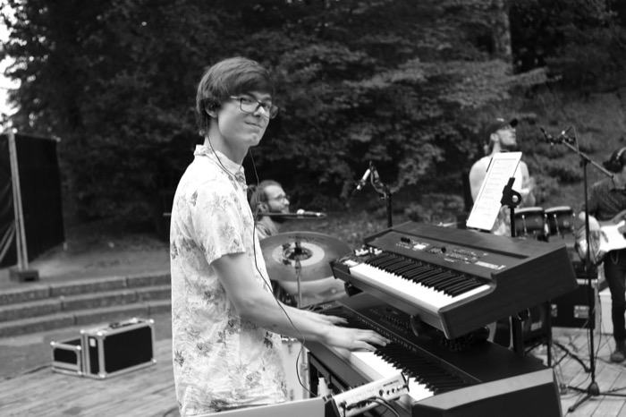 Keyboard player in a floral shirt glances over his shoulder mid-song at an outdoor stage, band behind him