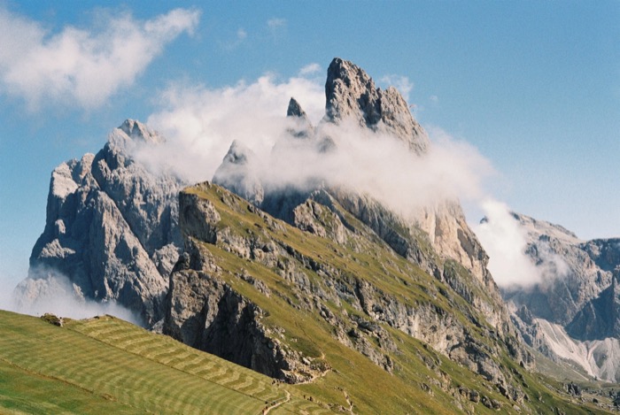 Cloud snagging on a serrated rock ridge above a green meadow slope cut with mowing lines