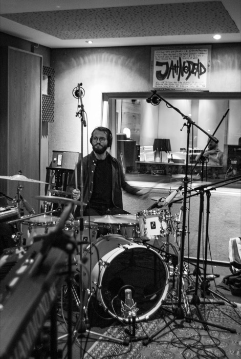 Bearded drummer in headphones behind a kit in a recording studio, hand-lettered poster above the control-room window