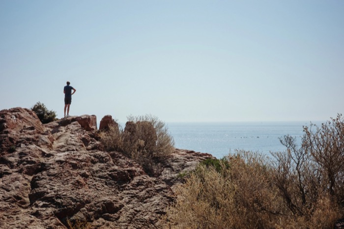 Small figure standing hands on hips atop a red rock outcrop, scrub and open sea beyond