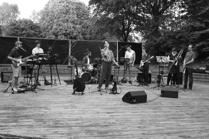 Eight-piece band playing on a wooden open-air stage in a park, singer at the microphone centre