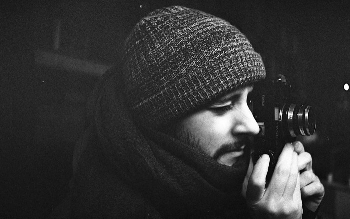 Man in a knitted beanie and scarf in profile, raising a rangefinder camera to his eye