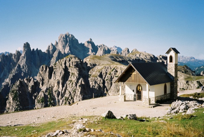 Small white mountain chapel with a bell tower on a gravel saddle, serrated rock spires rising behind