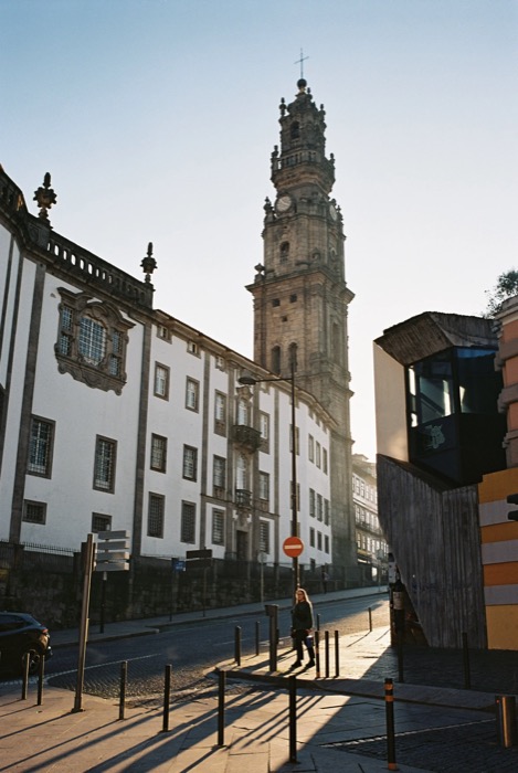 Woman walking past bollards below a tall baroque clock tower in low golden light