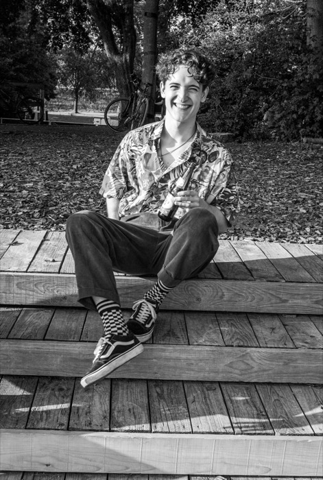 Young man in a patterned shirt and checkered socks laughing on wooden steps, beer bottle in hand