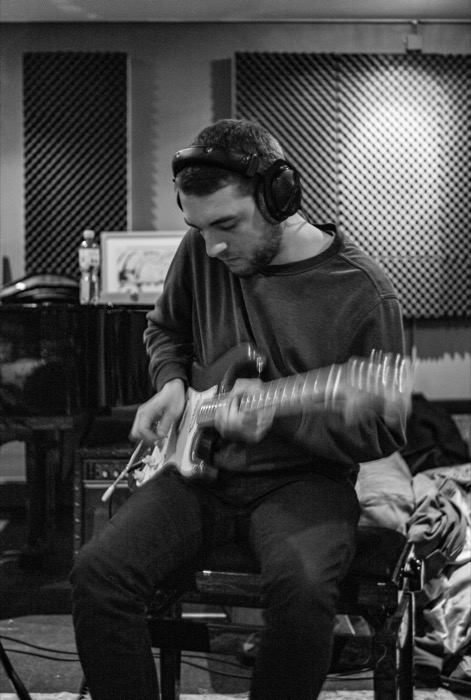 Young man in headphones playing an electric guitar in a foam-panelled recording studio, strumming hand blurred