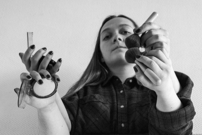 Woman holding a powder compact and makeup brushes up toward the camera, face behind her hands