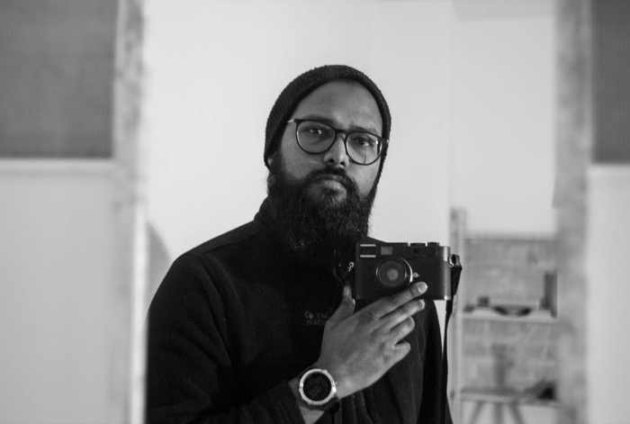 Bearded man in a knit beanie and glasses holding a rangefinder camera at chest height in a bare room