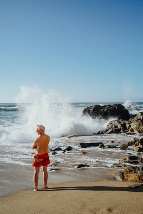 Man in red swim shorts standing on wet sand watching a wave burst white against the rocks