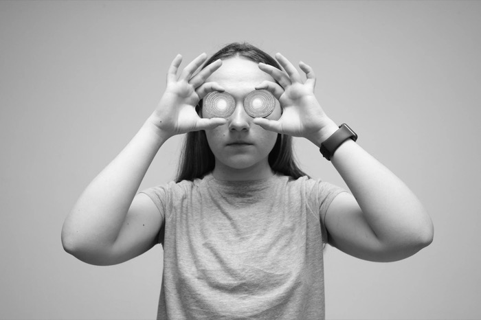 Girl holding two spiral-patterned discs over her eyes, hands framing her face against a plain wall