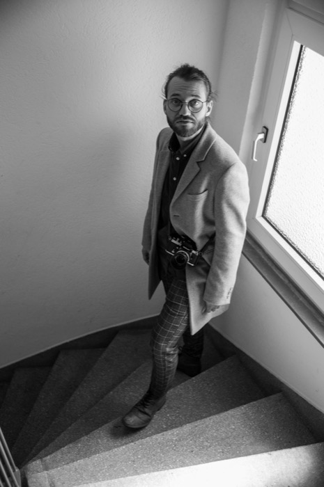 Man in a long coat with a film camera slung at his waist, pausing on a stairwell landing to look up