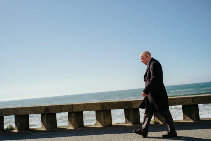 Elderly man in a dark overcoat walks head-down along a seafront promenade wall, waves behind him